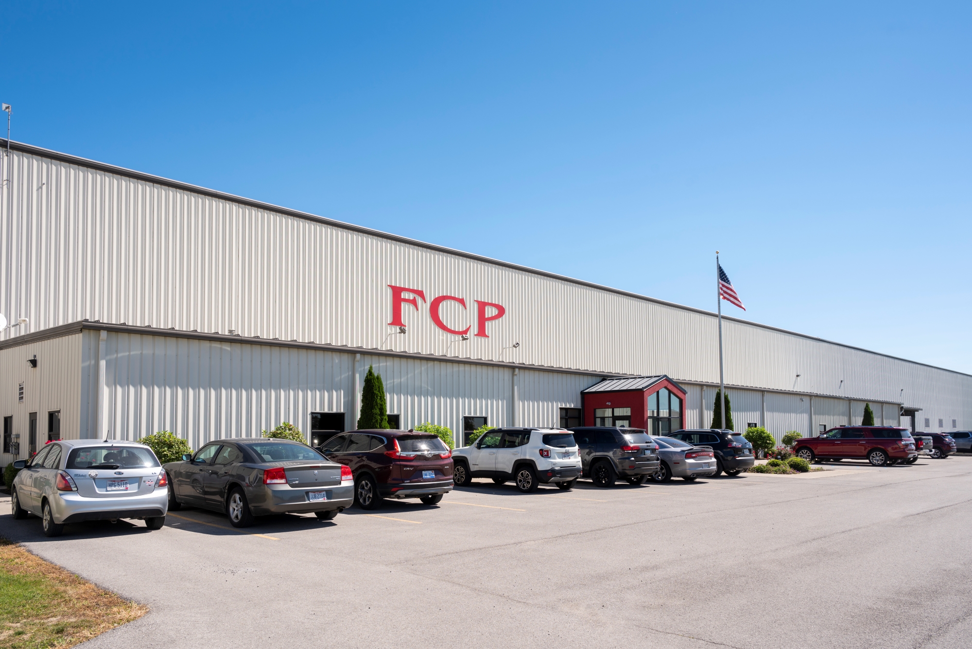 White industrial building with FCP signage, parking lot, cars, and flag under a clear blue sky.