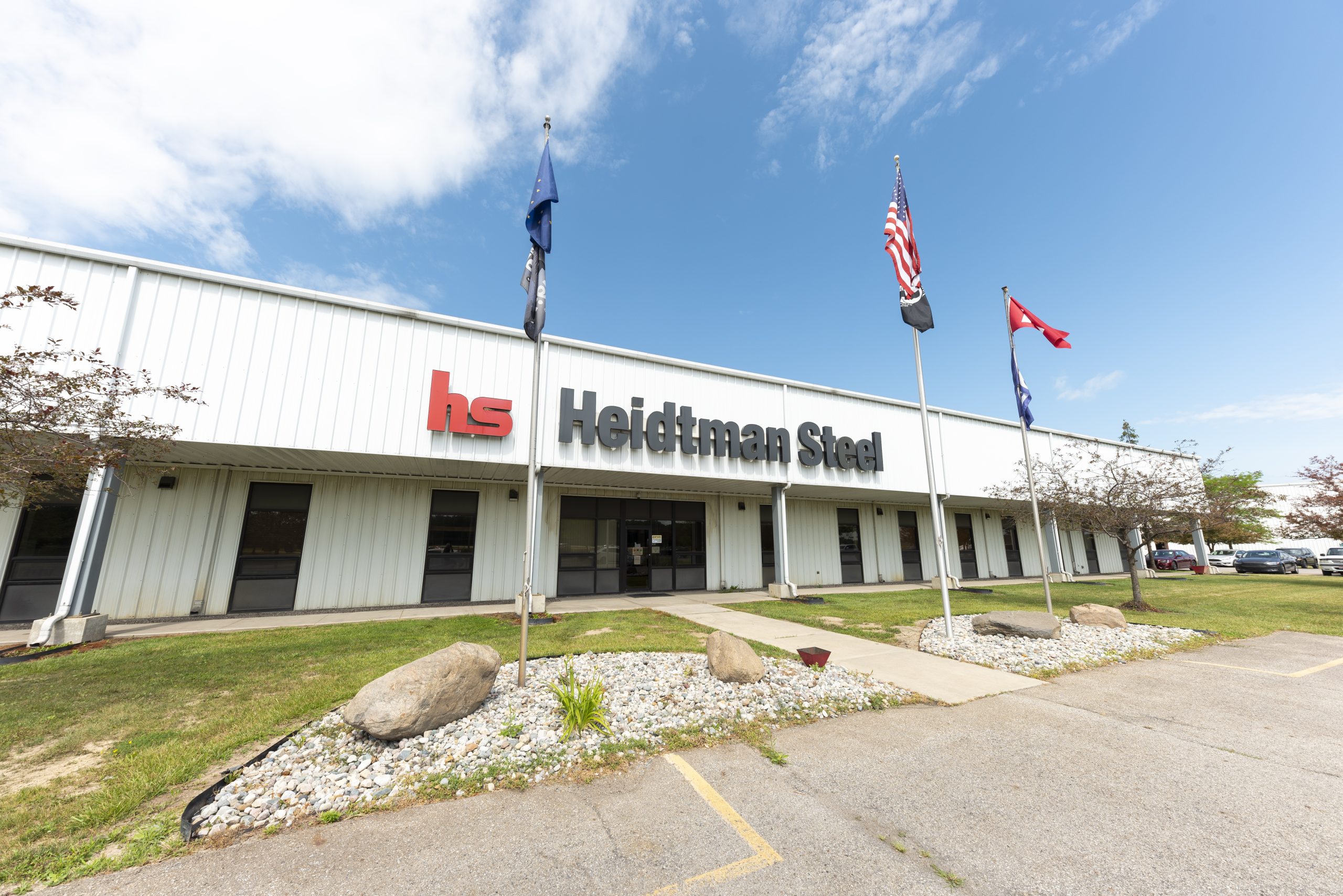 White industrial building with Heidtman Steel signage, flags, parking lot, and rocks under a blue sky.