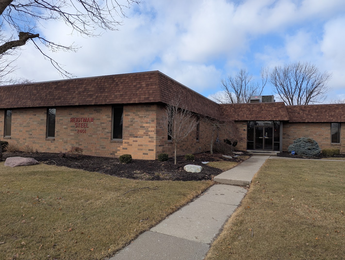 Brick office building with Heidtman Steel signage, walkway, and landscaping under a partly cloudy sky.