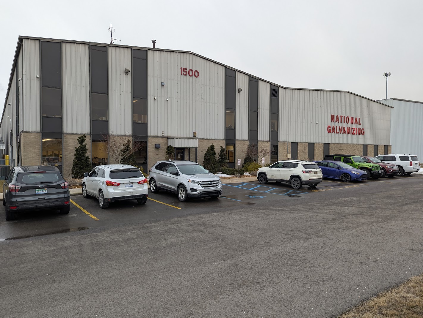 Gray industrial building with 'NATIONAL GALVANIZING' and '1500' signage, parking lot, and cars on a cloudy day.